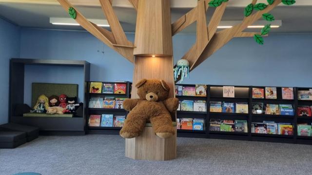 Interior of a library with a children's teddy bear sitting on a large wooden tree with wooden leaves