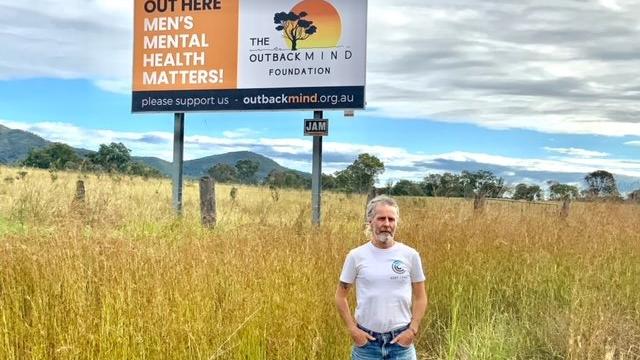 Man standing in a wheat field, in front of a large billboard advertising charity The Outback Mind