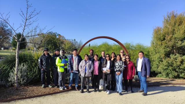 A group of 15 people standing outside on a sunny day in a botanical garden setting.