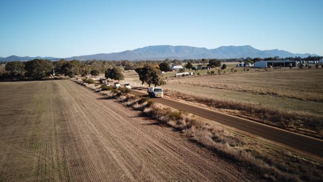 Very long shot of a rural road cutting through a paddock, with construction vehicles driving along it.