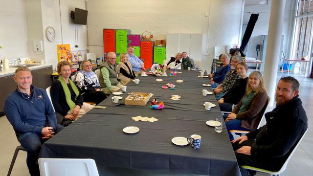 20 people sitting at a long table having morning tea, smiling at camera