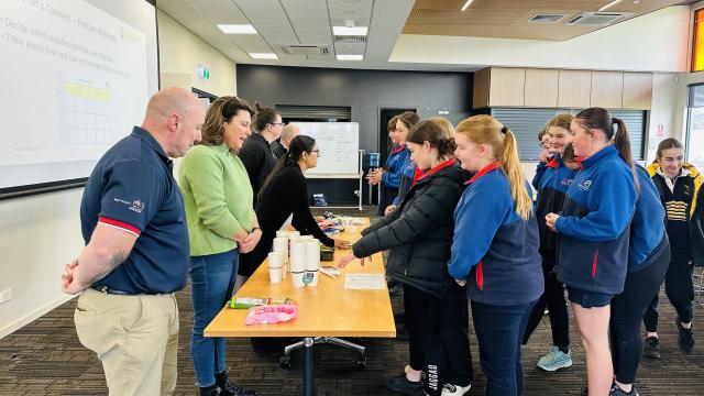A group of people; a trestle table in the middle of the room with a group of adults on one side and a group of engaged young students on the other