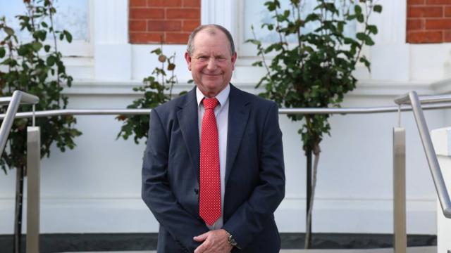A middle-aged man in a blue suit and bright tie smiling while standing in front of a building