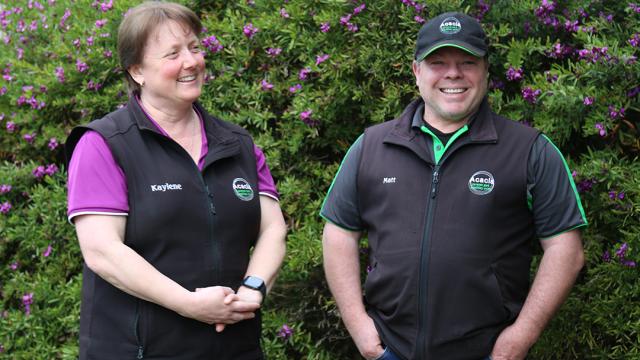 A man and a woman smiling and standing in front of a bush with purple flowers