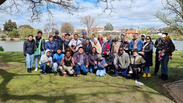 A large group of people standing in front of a lake in the sunshine, smiling for the camera