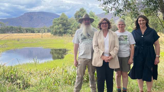 Four people stand together outdoors in front of a small pond, with trees and mountains in the background.