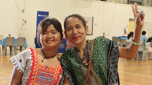 A young girl wearing traditional dress and a woman wearing a traditional print and red lipstick, smiling and leaning in together. The woman is making a peace sign with her left hand.