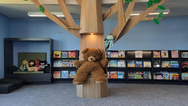 Interior of a library with a children's teddy bear sitting on a large wooden tree with wooden leaves