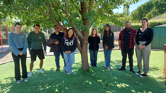Eight young people standing under a wide leafy tree on a sunny day and smiling at the camera
