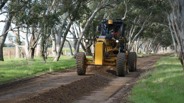 A grader is driving along a dirt road with green trees on either side