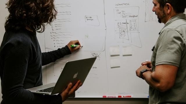 Two people collaborate at a whiteboard, with one person holding a laptop and writing on the board while the other watches, in a brainstorming or planning session.