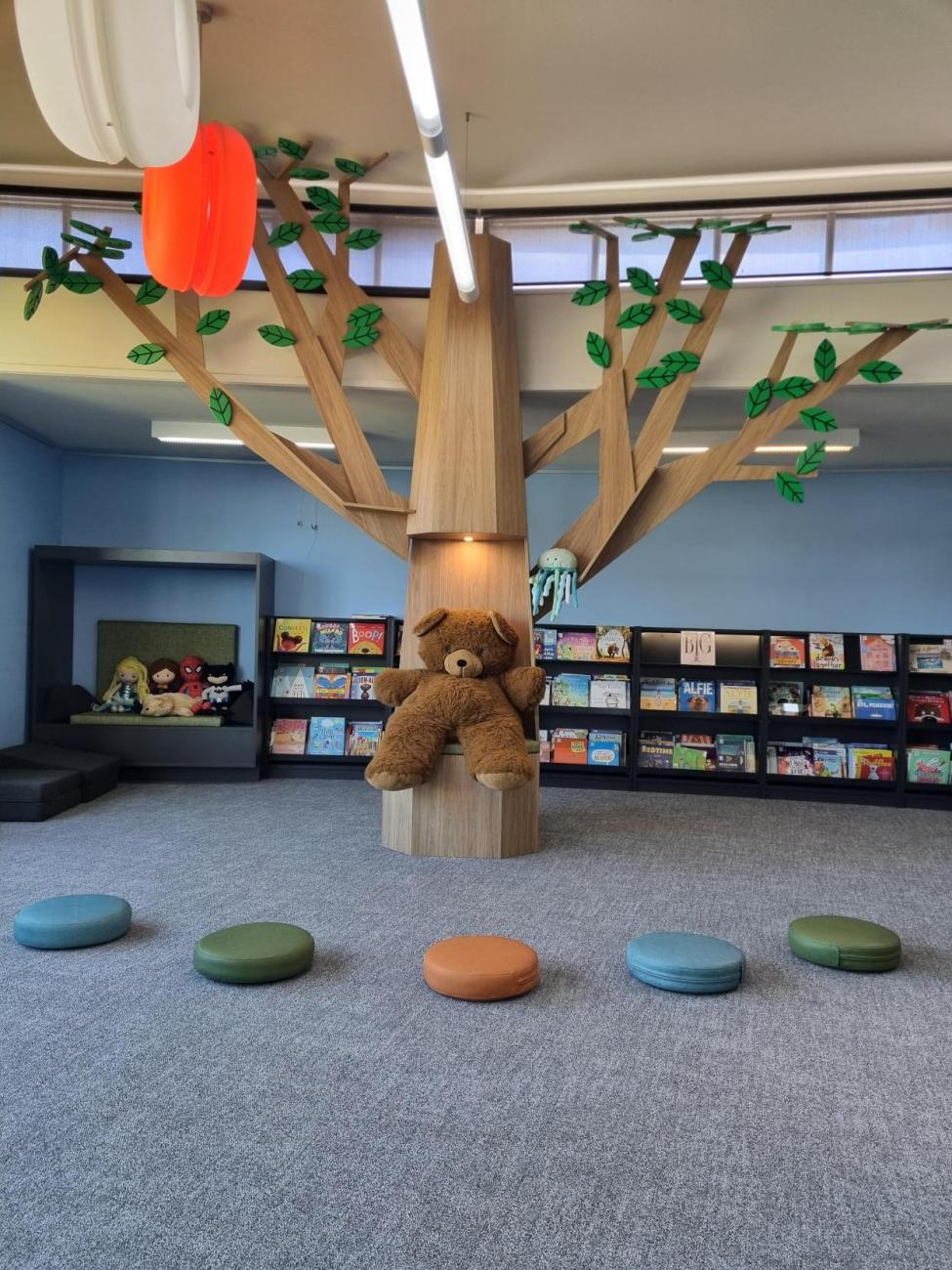 Interior of a library with a children's teddy bear sitting on a large wooden tree with wooden leaves