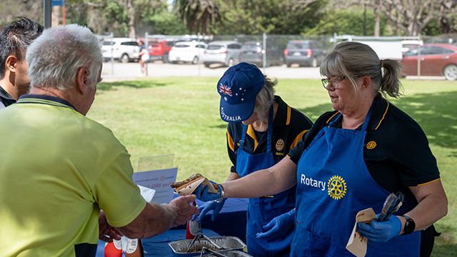 Rotary Club members cooking a sausage sizzle. A lady in a blue apron hands a sausage in bread to a white-haired man