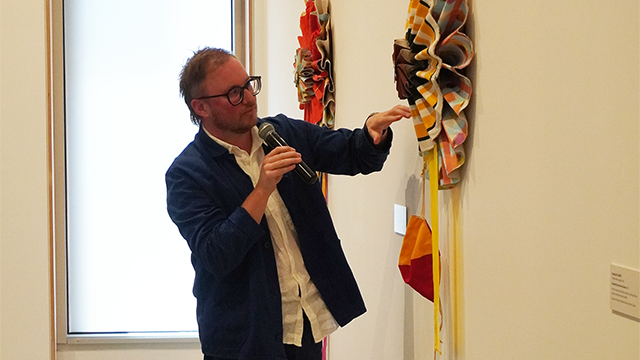 Young man in a blue suit speaking into a microphone while he gestures to an elaborate artwork mounted on a gallery wall