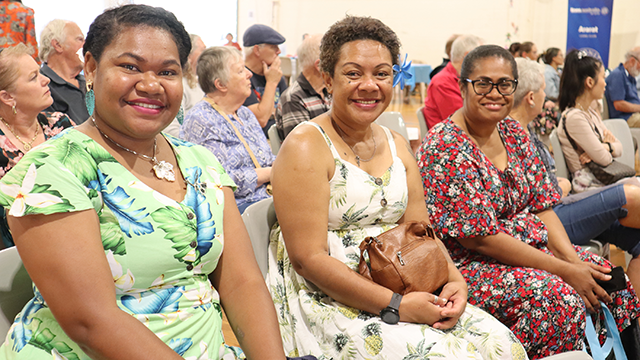 Three smiling women in brightly coloured dresses sitting in an audience of people, and smiling at the camera
