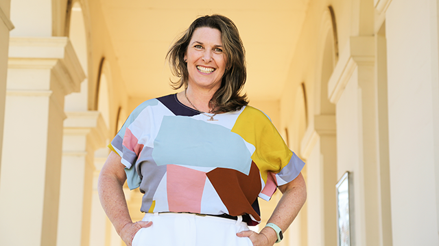 A middle-aged woman smiling for the camera, wearing a colourful blouse and with her hands in her pockets
