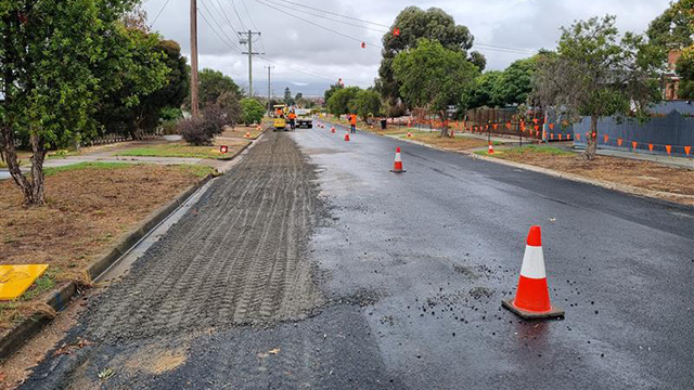 A residential asphalt road under construction with orange witches hats along the gravel edge