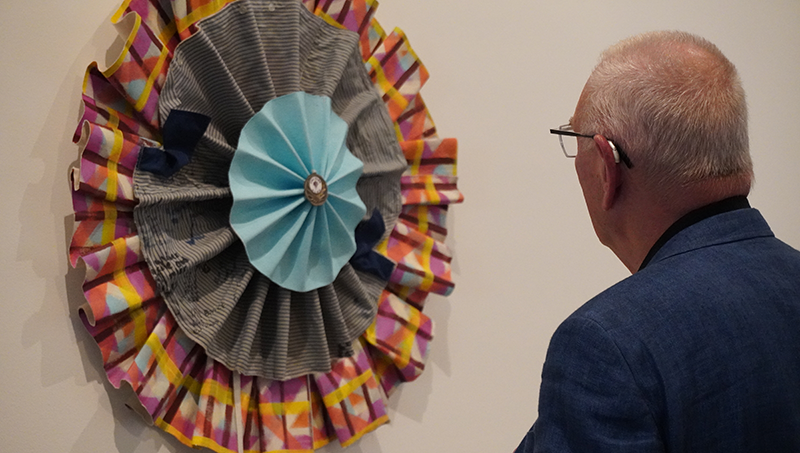 An older man looks closely at a textile artwork mounted on a gallery wall