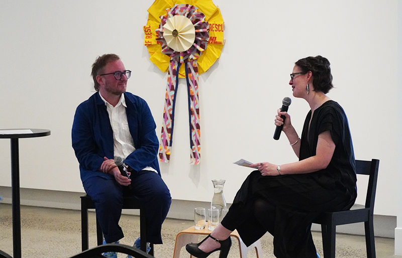 A man and a woman sit on stools in front of an audience. They are in conversation, each holding a microphone. The man is speaking and the woman is smiling at him. Behind them artworks hang on a gallery wall