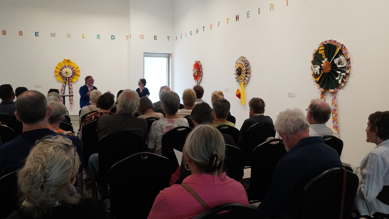 A gallery room full of people sitting on chairs and listening to a talk conducted by a young man in a blue suit