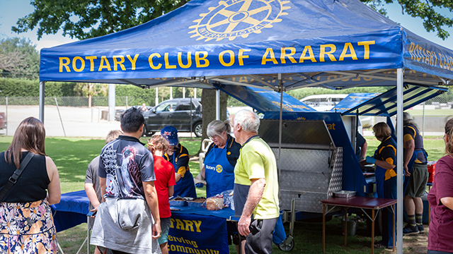 A group of people cooking sausages on an outdoor BBQ, underneath a bright blue and yellow marquee. People are living up to buy a sausage