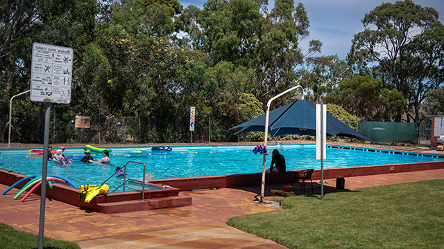 An outdoor pool with people playing and swimming, other sitting on lounge chairs, on a very sunny day