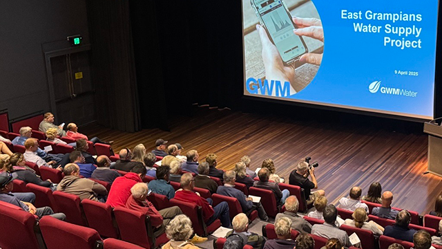 Long shot from the back of an auditorium with many people sitting in chairs looking at the presentation on a screen in front