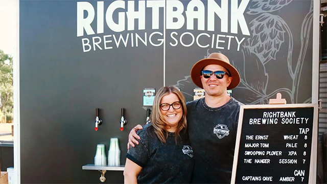 A man and a woman with their arms around one another's shoulders, standing behind an outdoor counter with a backdrop 'RIGHTBANK BREWING SOCIETY'